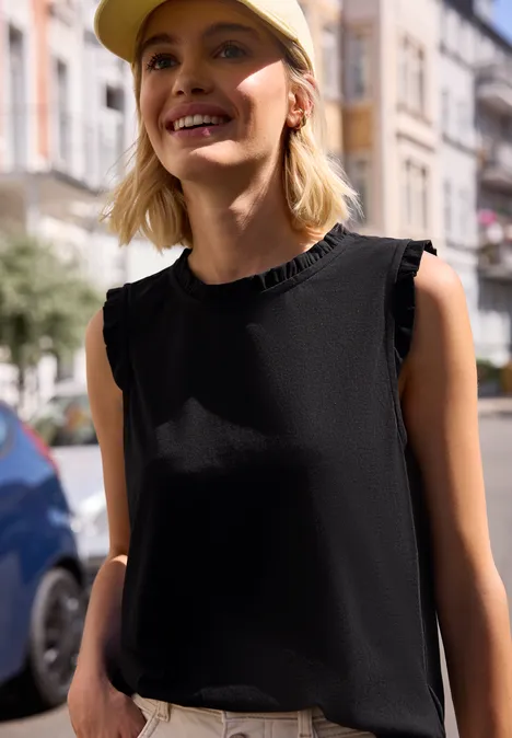 Een persoon draagt een zwarte mouwloze top met gerimpelde kraag en staat op een stadsstraat.